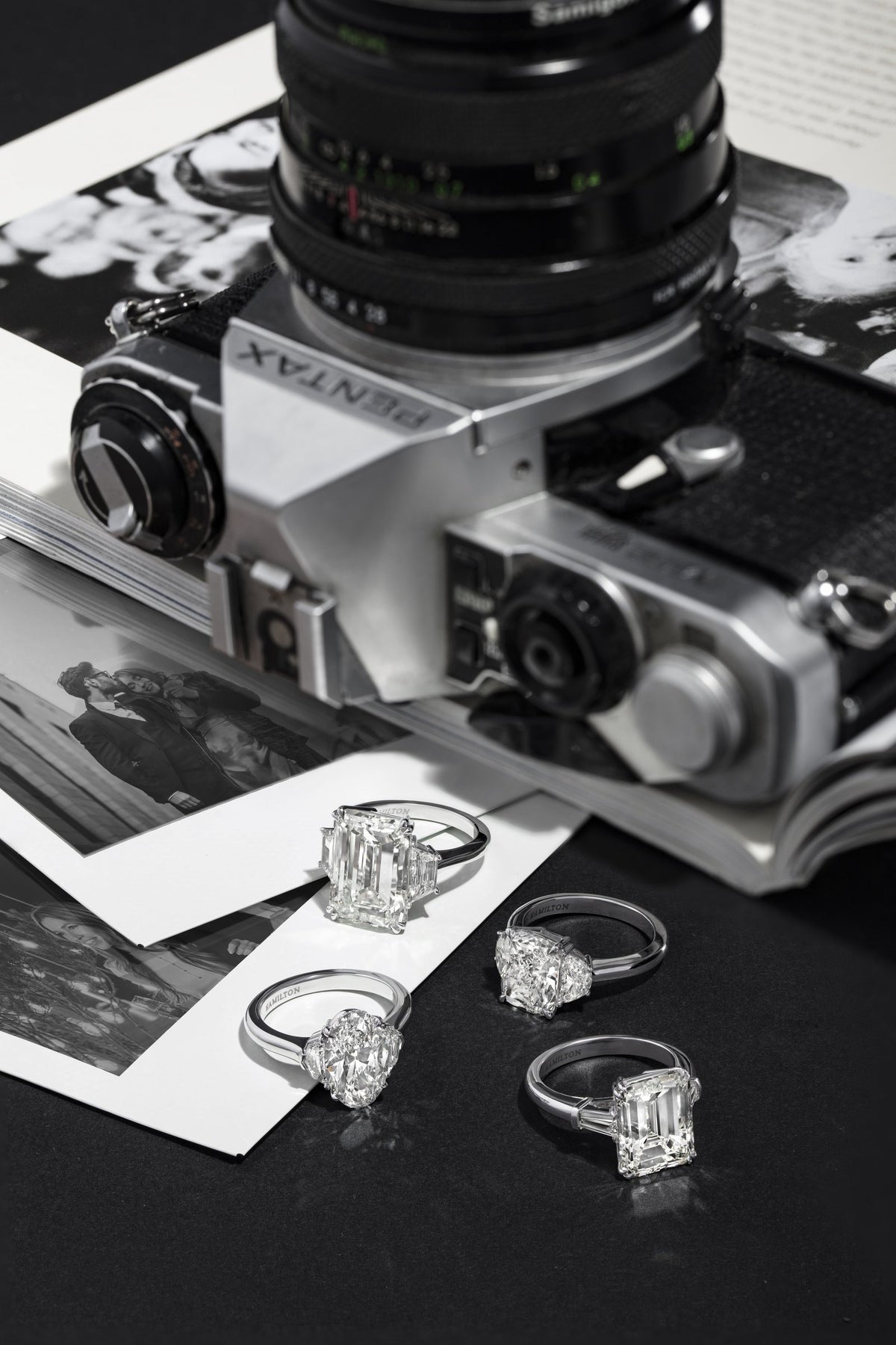 Four diamond solitaire rings in emerald, pear, oval and round cuts arranged beside a vintage film camera on black-and-white photographs