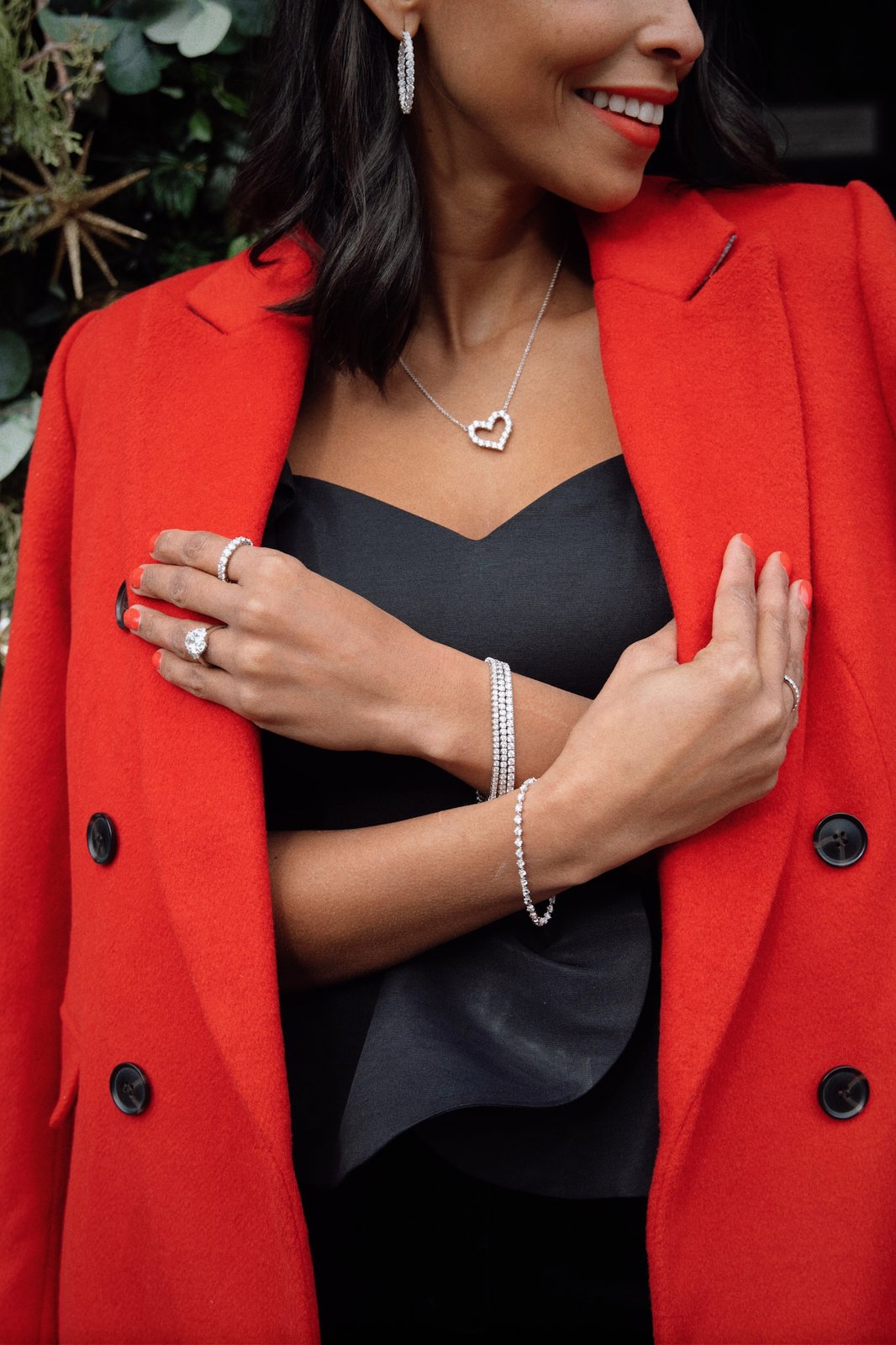 Smiling woman in a red coat wearing a diamond heart pendant, diamond hoops, a tennis bracelet and diamond rings