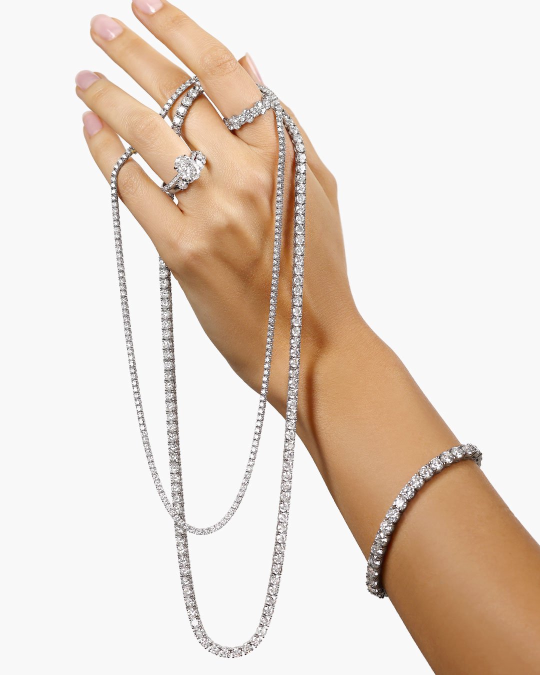 Hand draped with diamond tennis necklaces, a diamond tennis bracelet and diamond rings on a white background