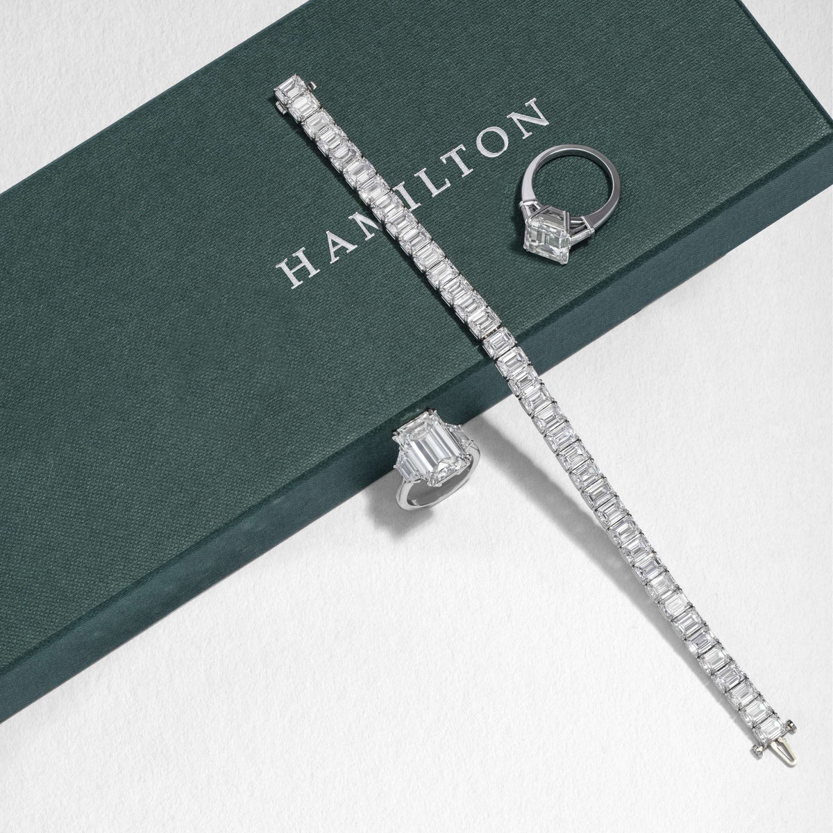 Emerald-cut diamond tennis bracelet and two emerald-cut diamond rings on a green Hamilton Jewelers box