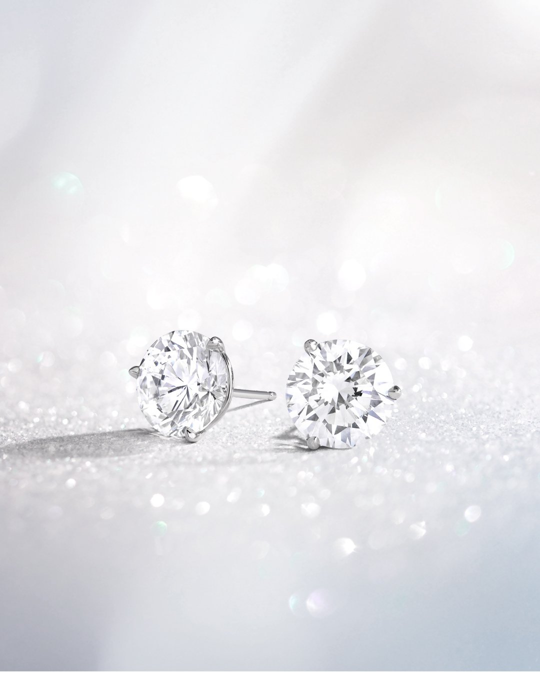 Pair of round brilliant diamond stud earrings in three-prong martini settings on a sparkling white surface