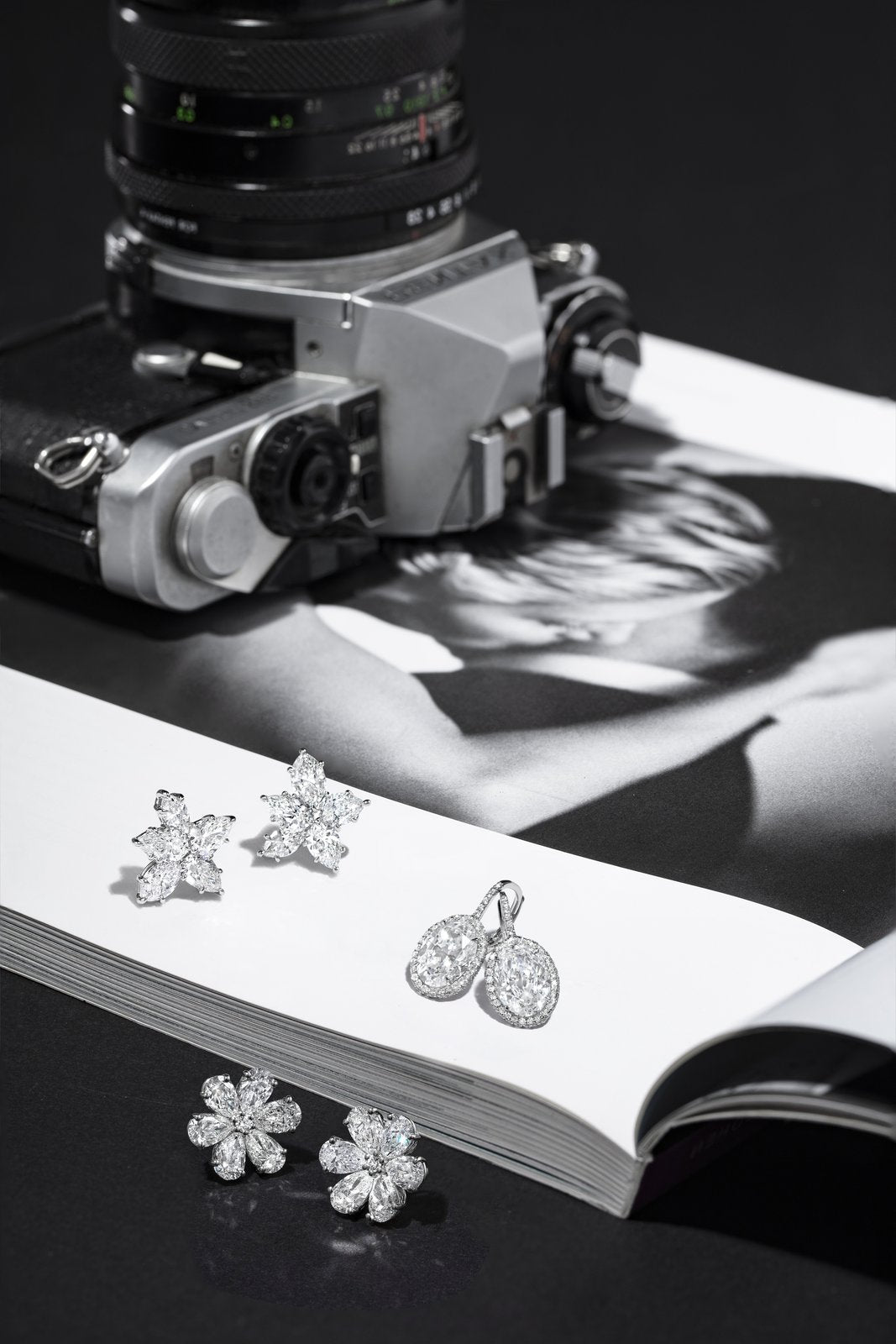 Diamond flower cluster earrings and oval drop earrings laid on an open magazine beside a vintage camera