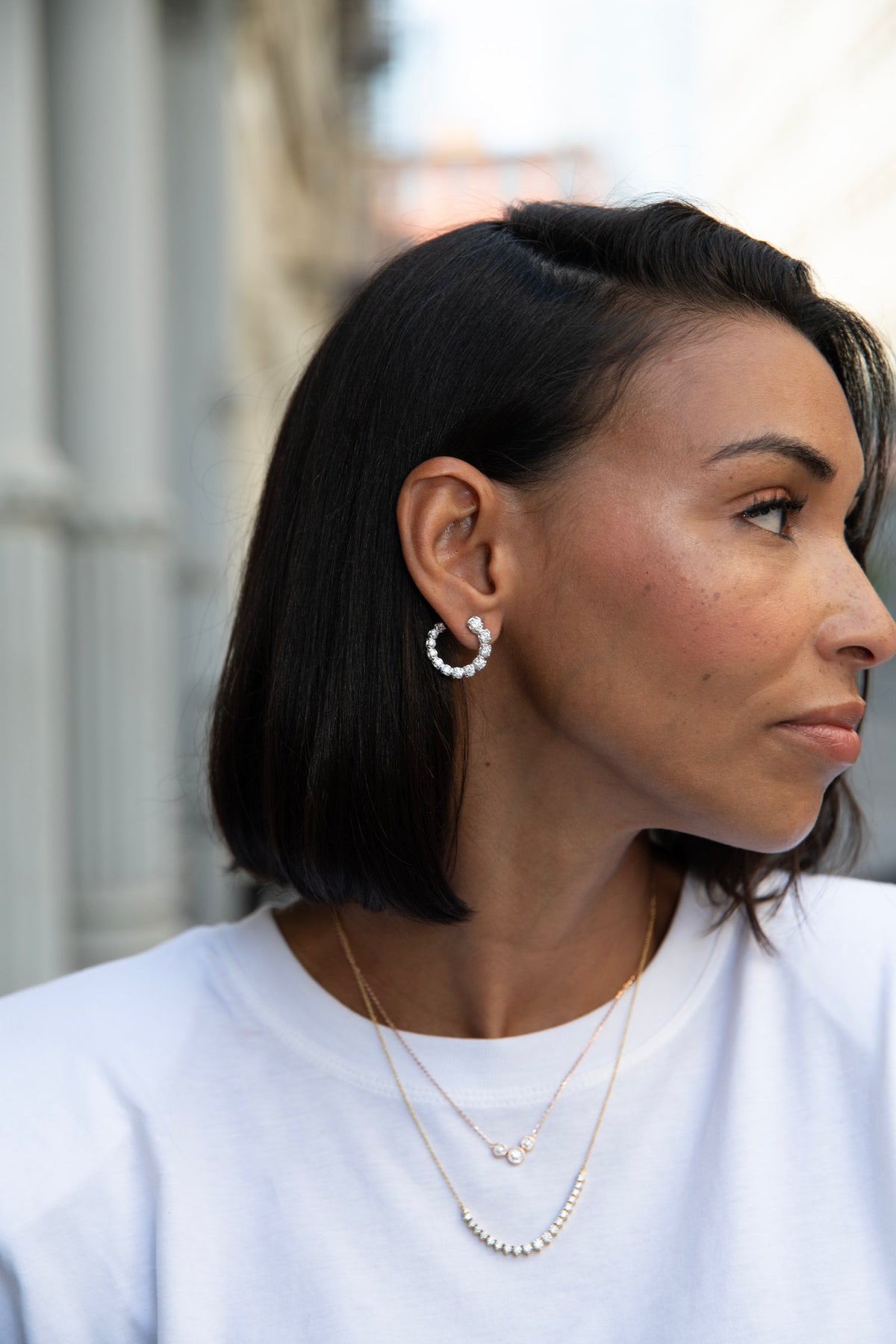 Profile of a woman with a dark bob wearing a diamond inside-out hoop earring and layered diamond necklaces on a city street