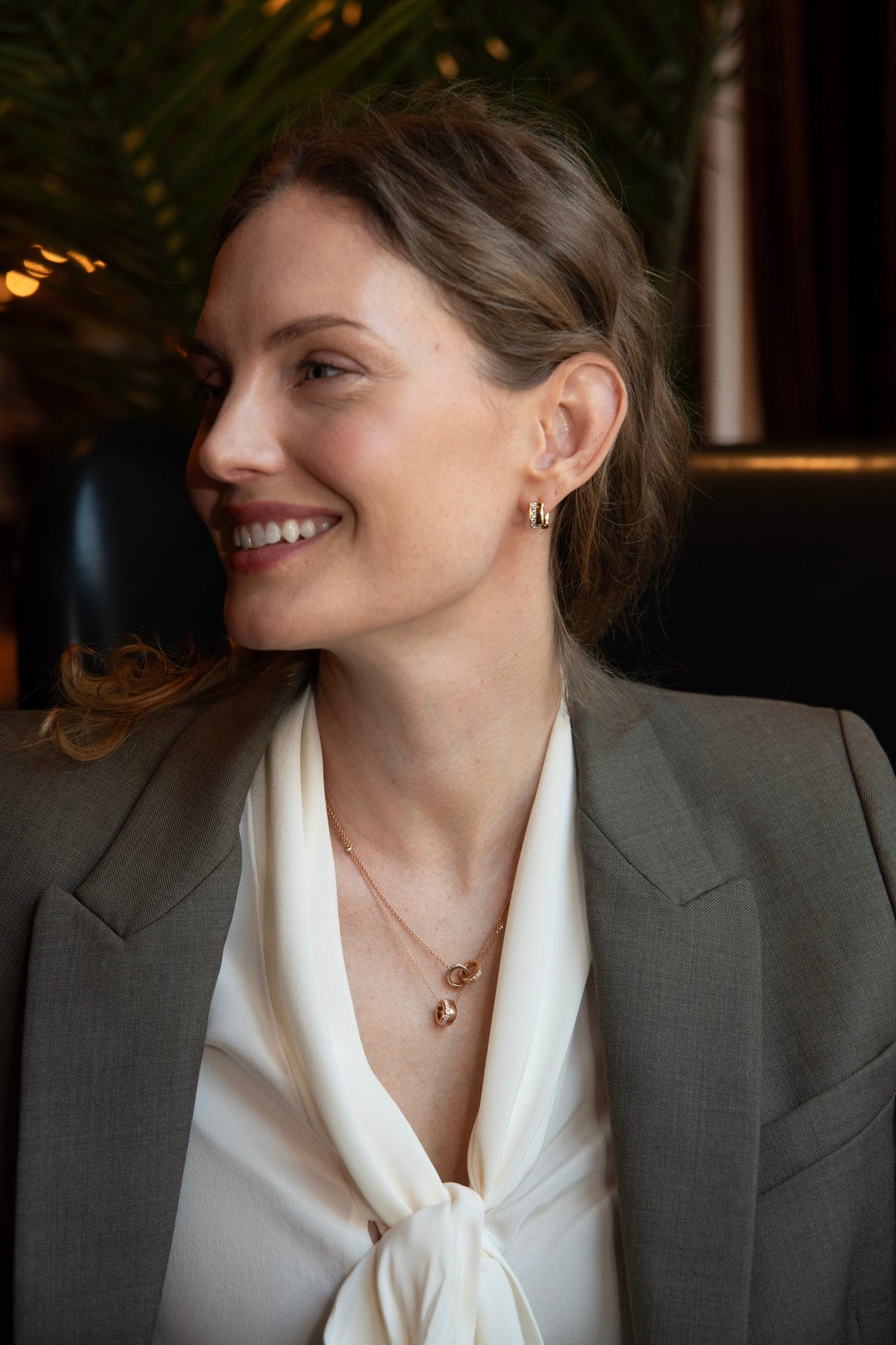 Smiling woman in a gray blazer and white blouse wearing a delicate gold Eternity necklace and small hoops