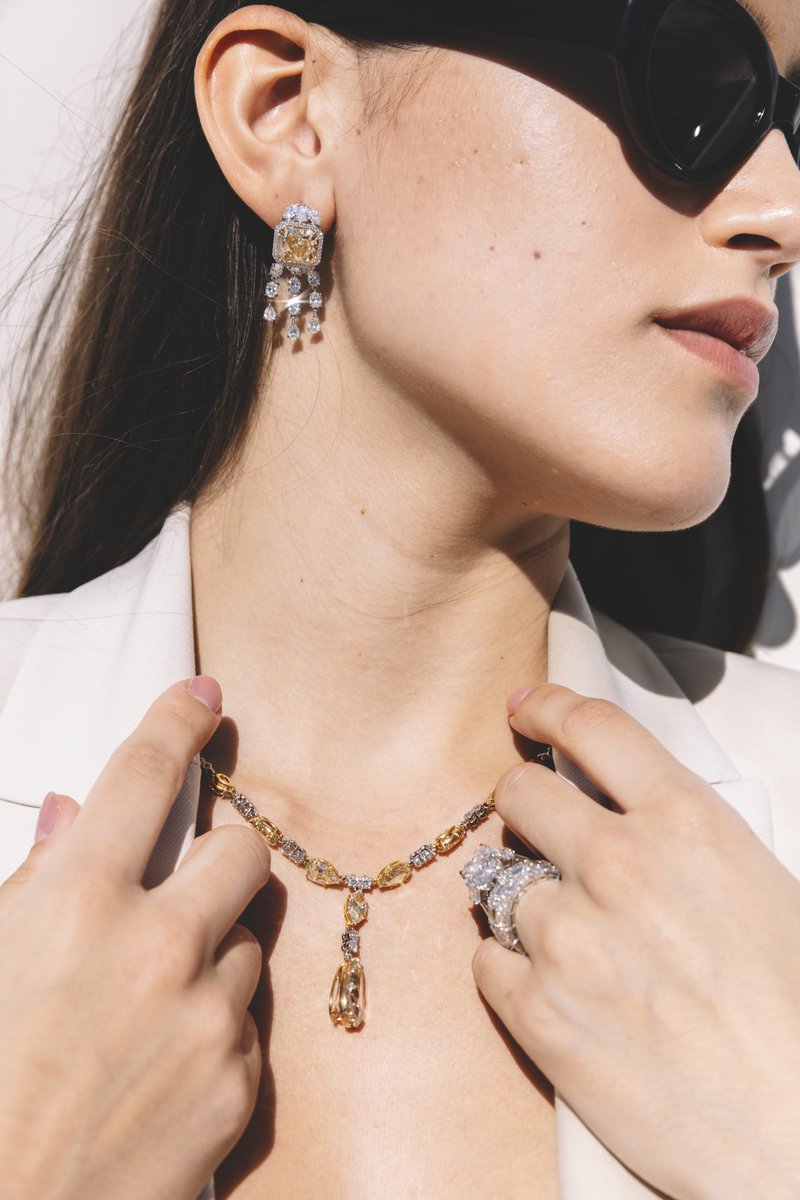 Woman in sunglasses and a white blazer holding a multicolor gemstone necklace with a drop pendant, wearing diamond earrings